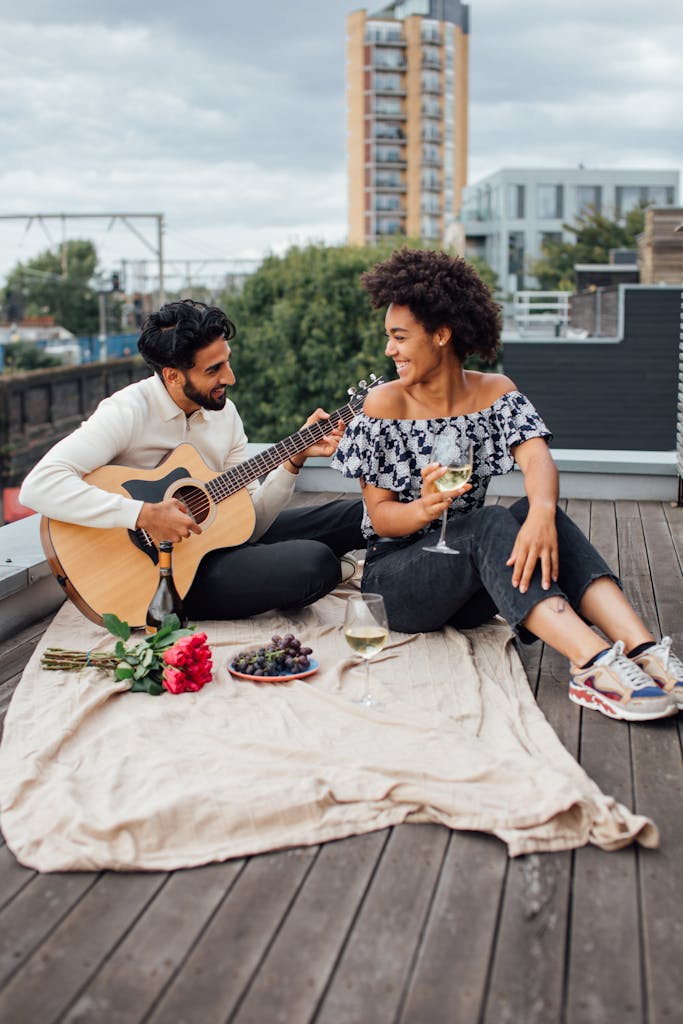 A couple enjoys a romantic rooftop picnic with wine and guitar music, captured in a vibrant outdoor setting.