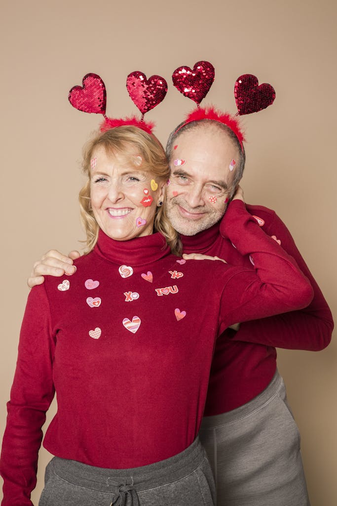 Happy senior couple celebrating Valentine's Day with heart accessories and red sweaters, embracing lovingly.